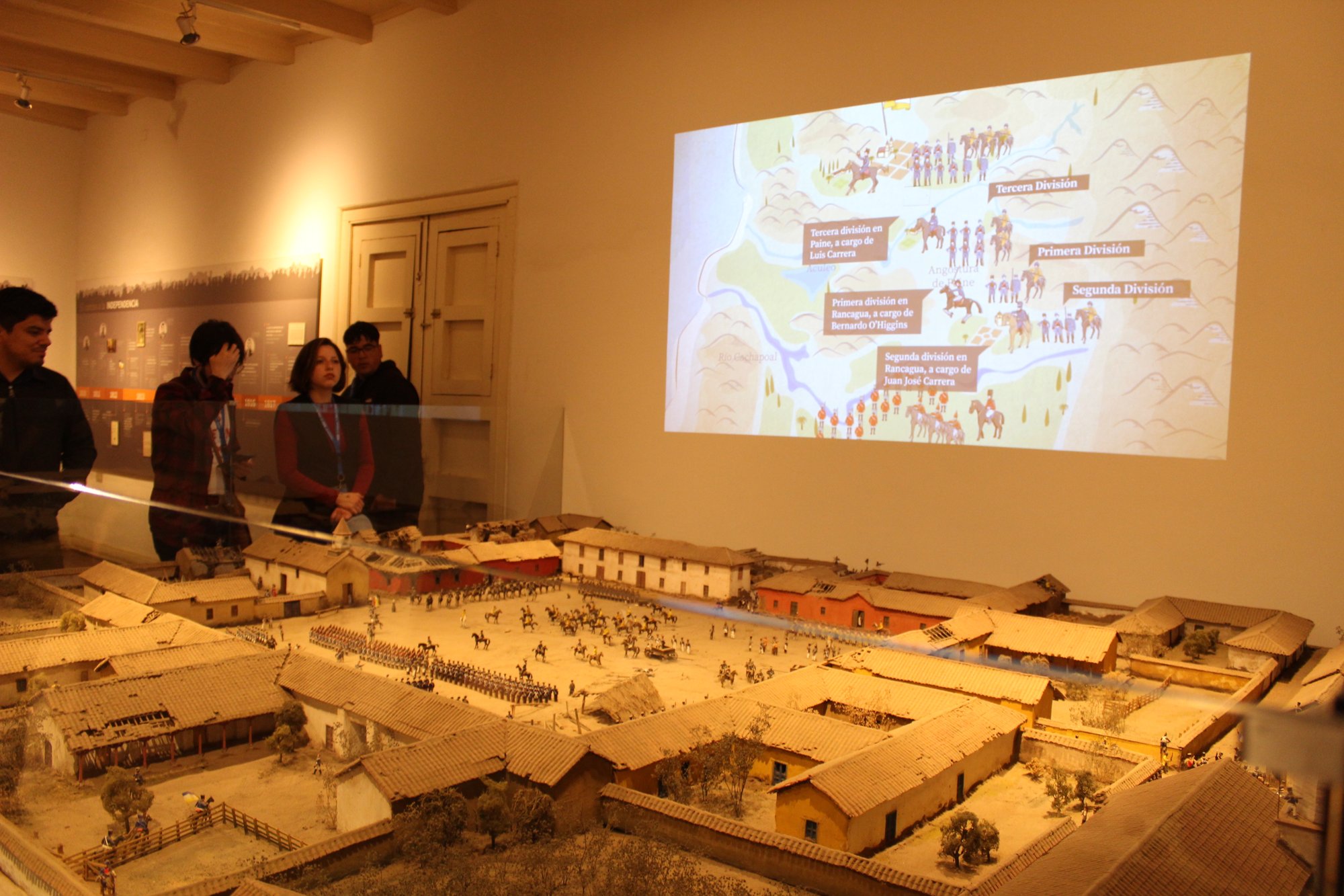 Varias personas observan una maqueta junto a una proyección en una sala de exposición.