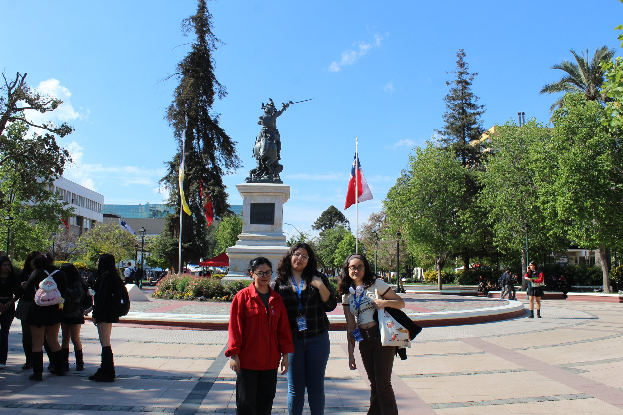 Tres personas posan en una plaza con árboles y un monumento al fondo.