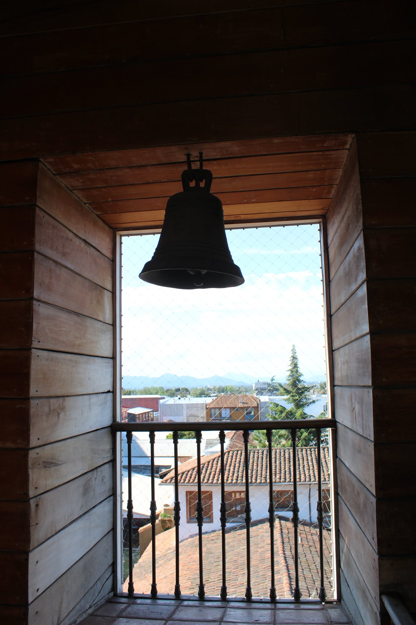 Una campana cuelga frente a una abertura con vista a los tejados.