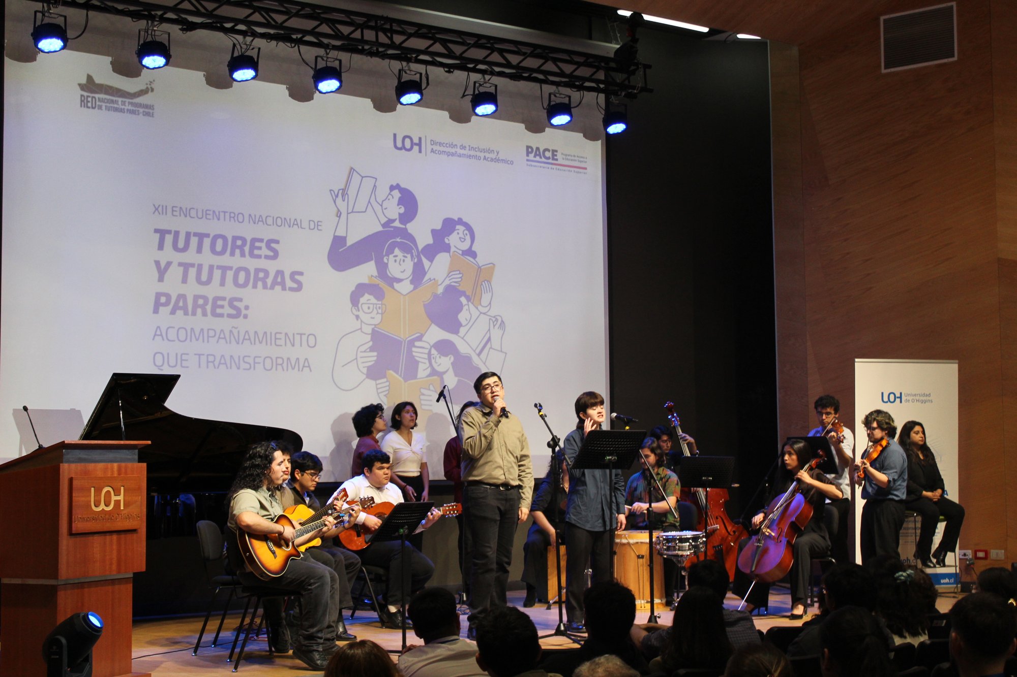Personas con guitarras y otros instrumentos se reúnen en el escenario del auditorio.