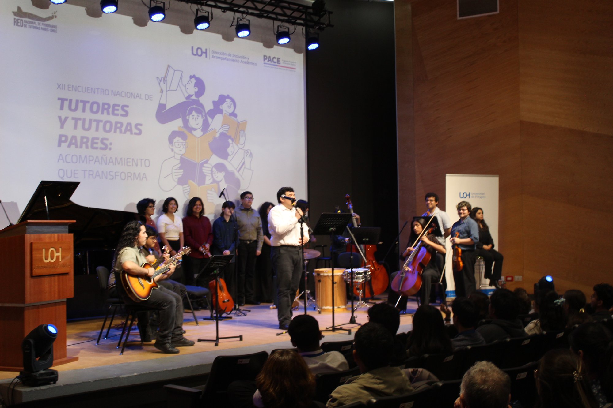 Un conjunto musical ocupa el escenario del auditorio, con público en primer plano.