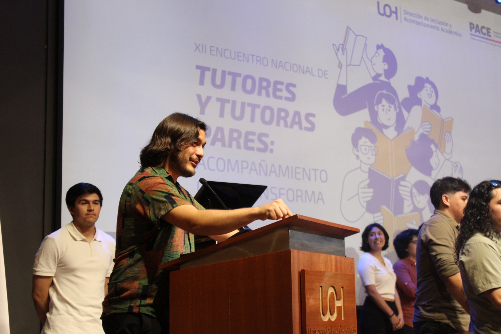 Una persona habla al micrófono desde un atril, con otras personas detrás.