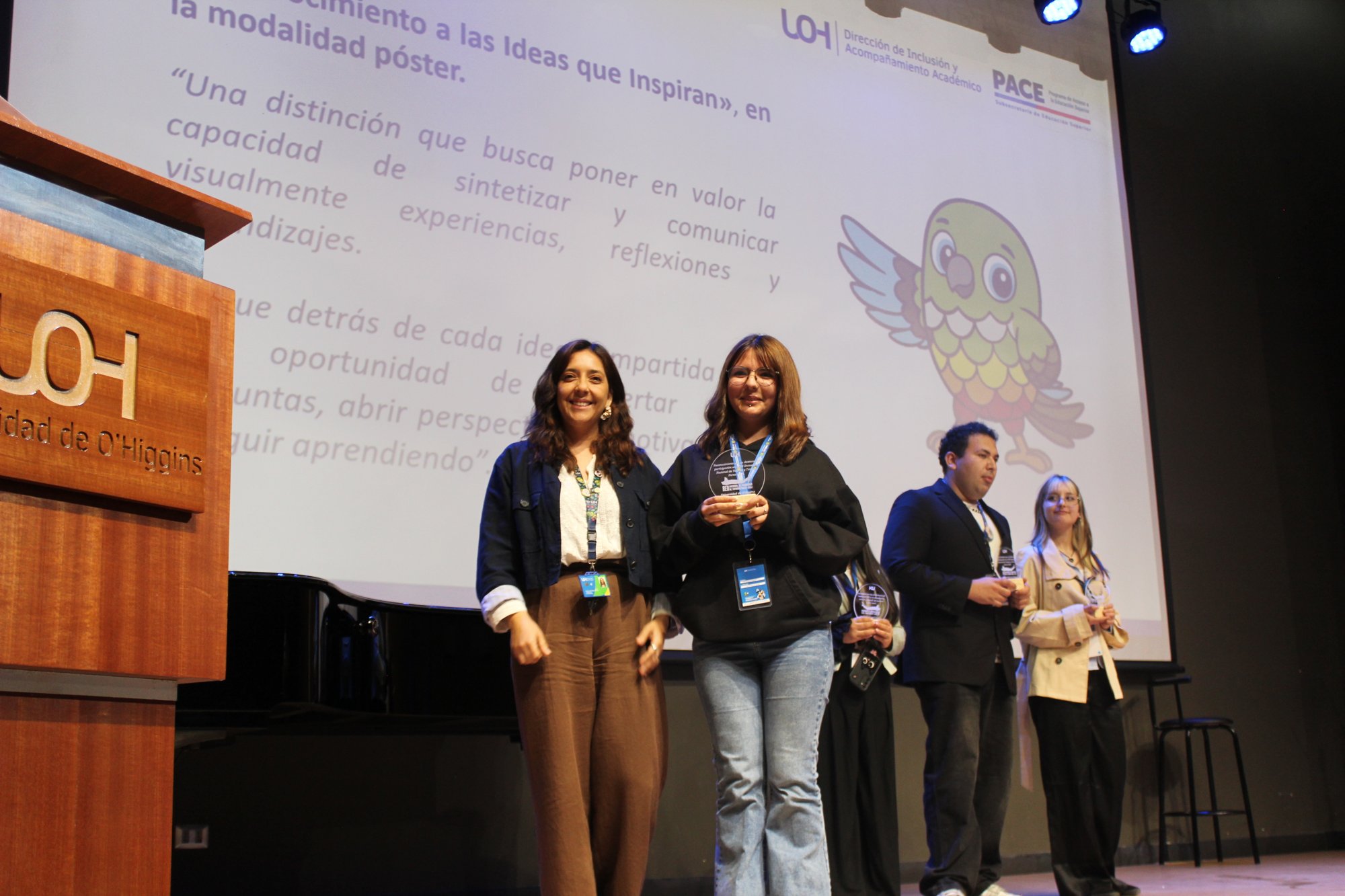 Dos personas posan en primer plano sobre el escenario, con otras personas al fondo.