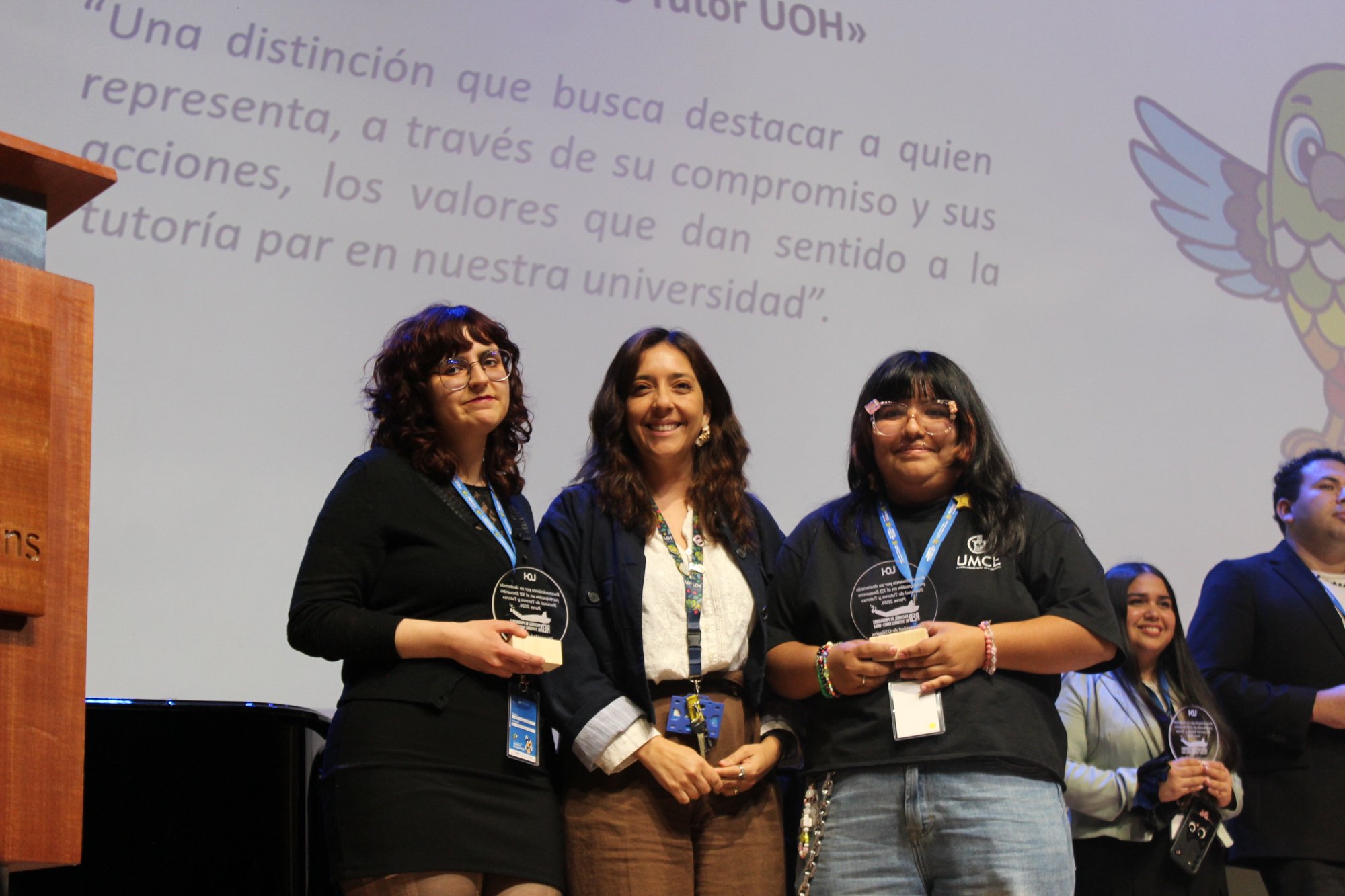 Tres personas posan juntas sobre el escenario frente a la pantalla del encuentro.